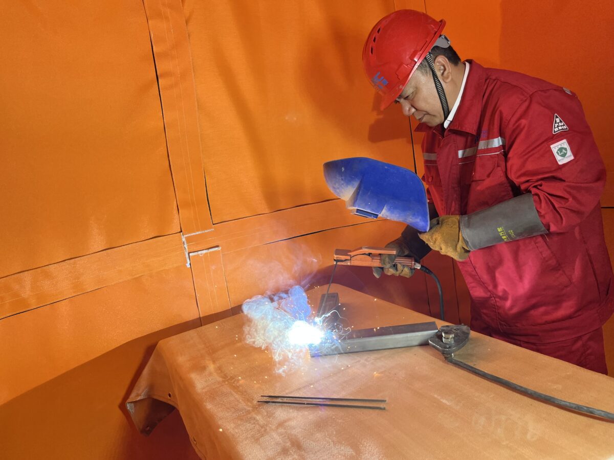 Welder performing hot work inside a pressurized welding enclosure at an industrial plant, with welding sparks contained by flame-retardant panels and positive-pressure ventilation equipment.
