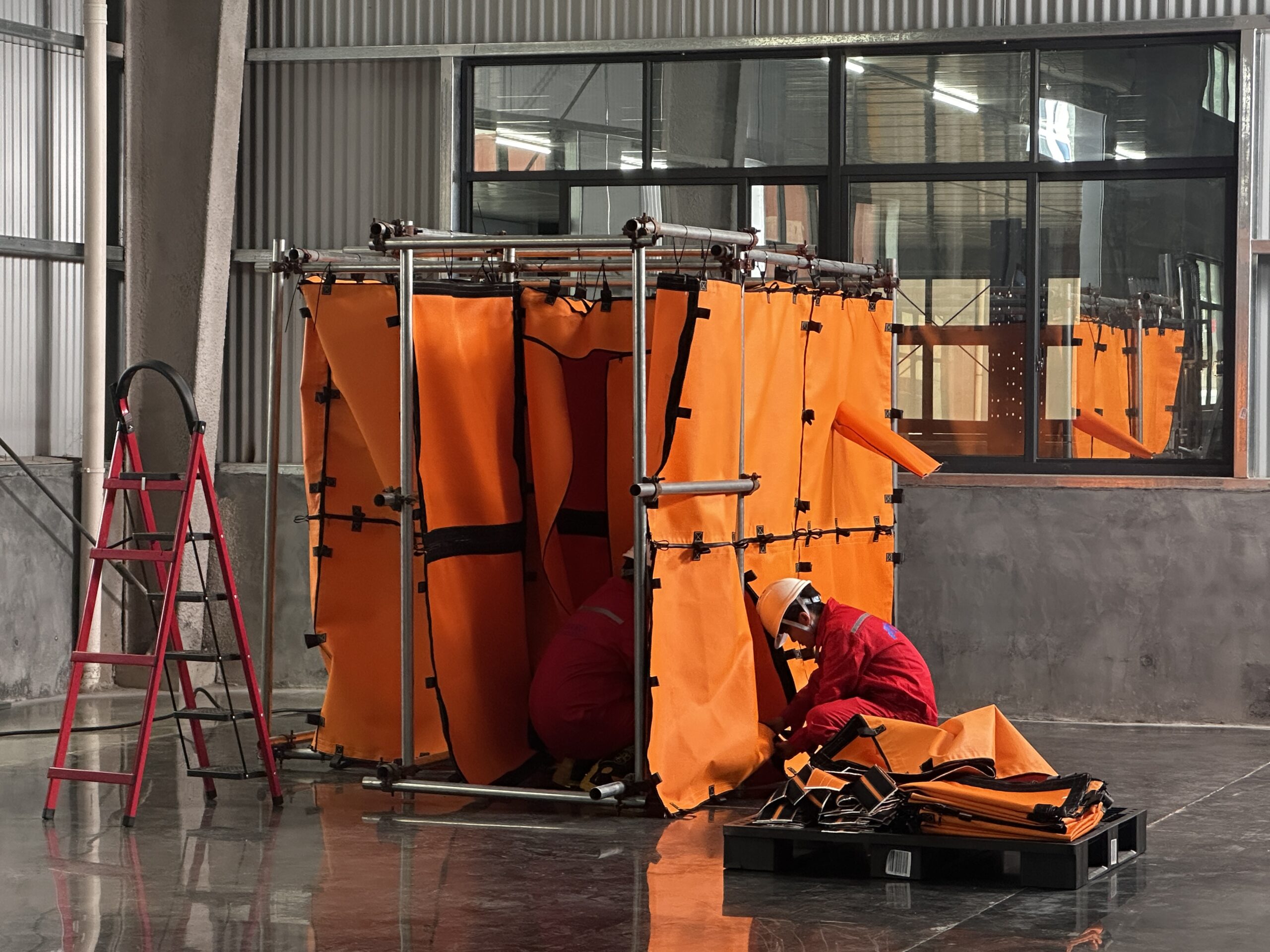 Workers installing an orange fire-resistant welding habitat system with modular frame and protective fabric panels in an industrial workshop.